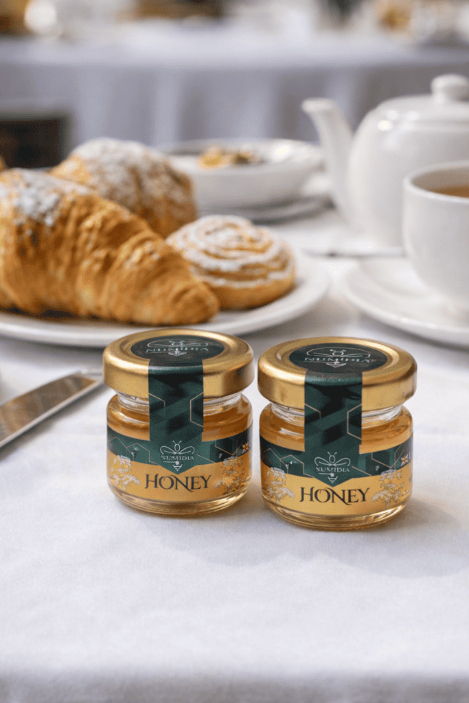 Two 28g Numidia mini honey jars on a restaurant breakfast table beside croissants, pastries, and tea, showcasing premium local honey for guests.